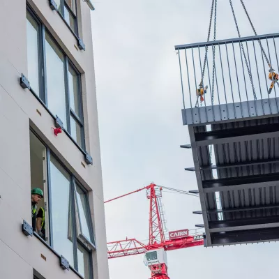 Prefabricated balcony being lifted by crane next to a multi-storey building under construction.