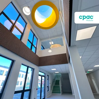 A bright, modern school interior with large windows, a central skylight featuring a yellow circular frame, and a staircase leading to the upper floor.