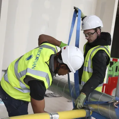 Two construction workers in high vis jackets, white hard hats, and goggles work on tying a pipe